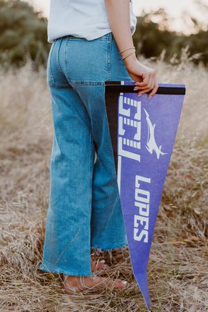 Person holding a purple GCU Lopes pennant flag in a field wearing blue flare jeans.