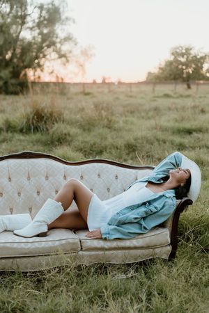 Woman in denim jacket and white boots reclines on vintage sofa outdoors at sunset in a grassy field.