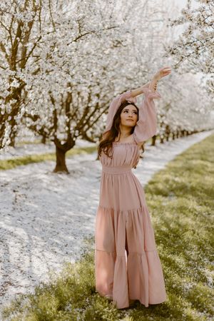 Woman in pink maxi dress reaching toward white blossoming trees in a serene spring orchard pathway.