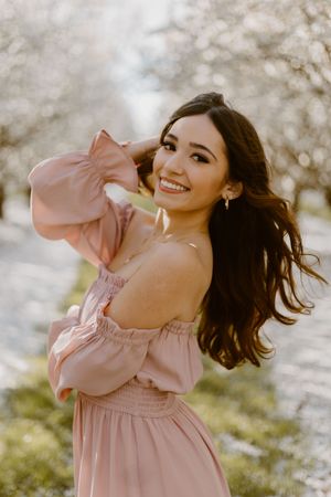 A smiling young woman in a pink off-shoulder dress poses among blooming white trees in a spring orchard.
