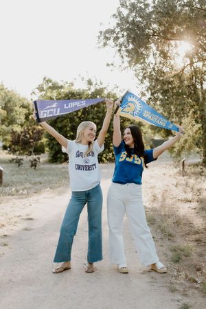Two college women hold GCU Lopes and SJSU Spartans pennants outdoors on a sunny dirt path.