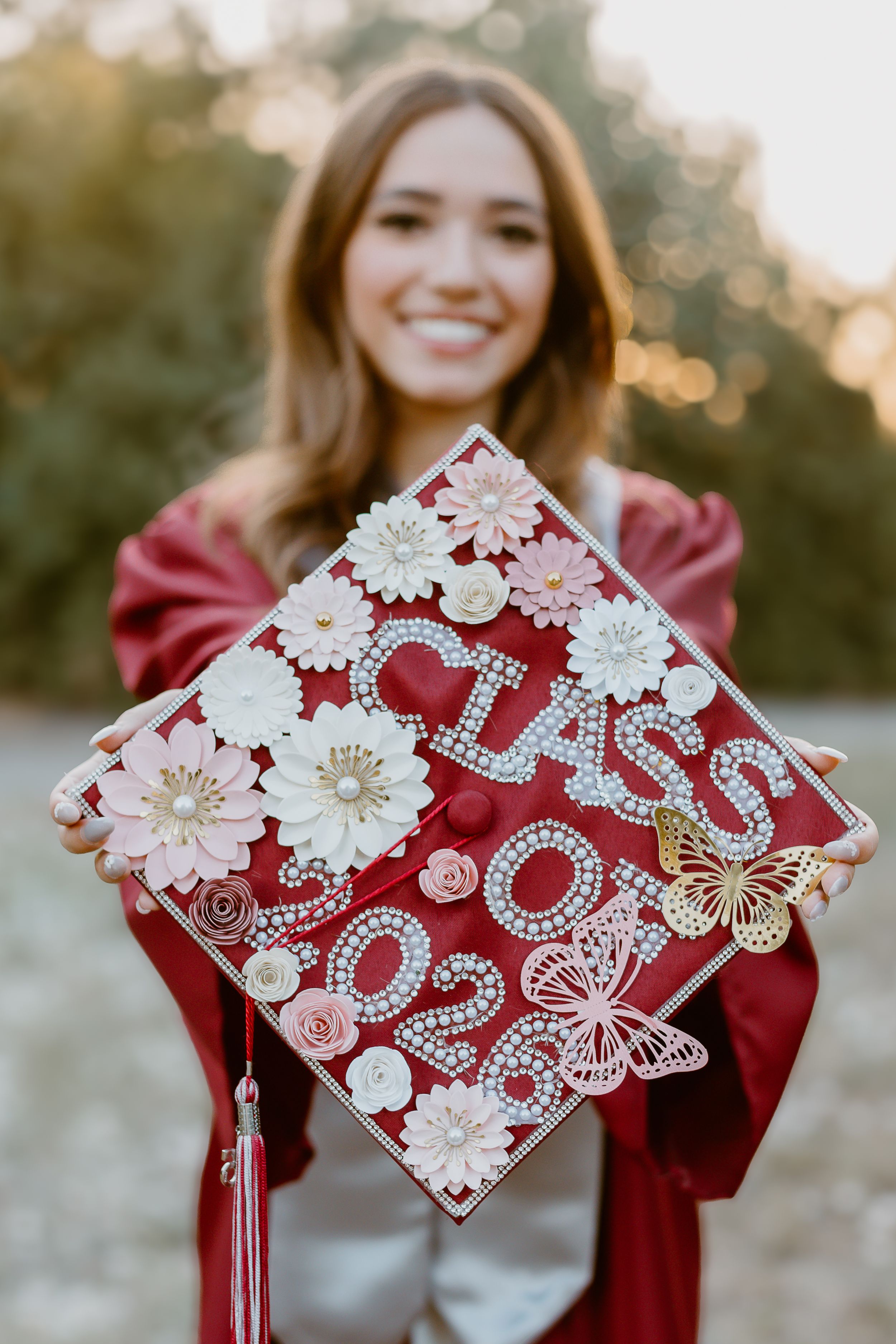 Smiling graduate in red gown holds decorated Class of 2023 cap with flowers, rhinestones, and butterfly.