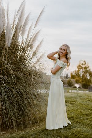 A young woman in a white pleated dress poses gracefully beside tall pampas grass in a serene outdoor setting.