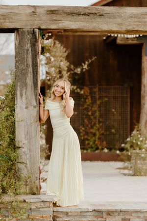 Smiling blonde woman in a pale yellow off-shoulder maxi dress posing by a rustic wooden pergola outdoors.