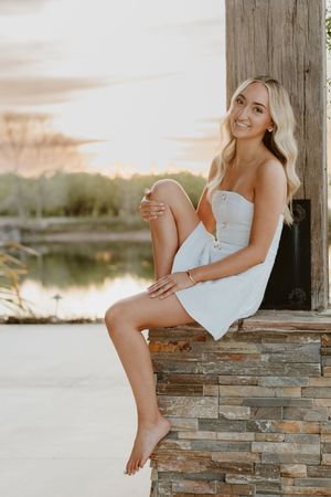 Young blonde woman in white strapless dress sitting on stone ledge near waterfront at sunset.