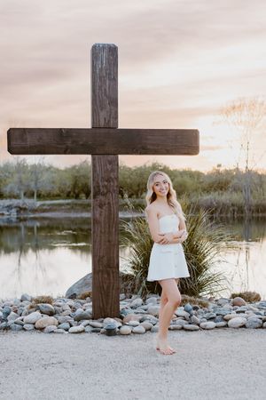 Blonde woman in white outfit standing barefoot beside a wooden cross near a serene lake at sunset.