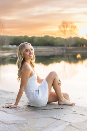 Smiling blonde woman in white dress sitting on stone by a reflective lake at golden sunset.