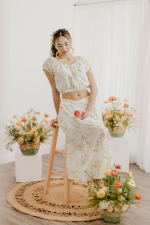 Woman in floral crop top and maxi skirt sitting on wooden stool surrounded by colorful flower arrangements.