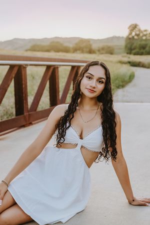 Woman in white dress poses on wooden bridge during sunset with mountains and greenery in background.