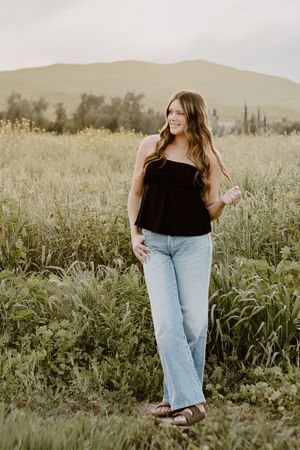 Young woman in black top and light jeans standing in a lush green meadow with rolling hills in the background.