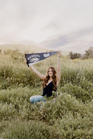 Young woman sitting in a green meadow holding a University of Nevada pennant flag raised above her head.