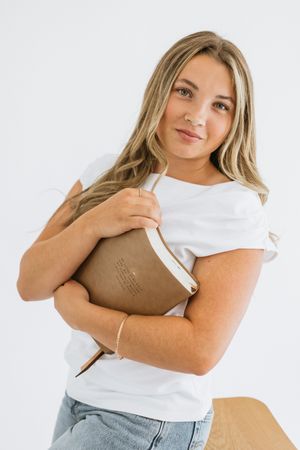 Young woman in white t-shirt and jeans hugging a brown leather notebook, smiling against white background.