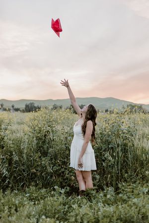 Young woman in white dress releasing a red heart balloon in a lush green field at sunset with mountains in background.