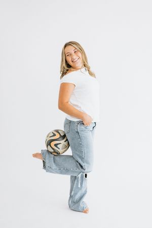 Smiling blonde woman in white tee and wide-leg jeans balancing a soccer ball on her foot against white background.