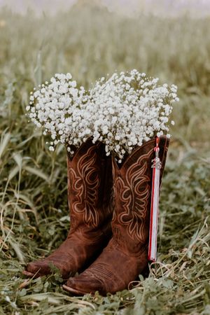 Brown cowboy boots filled with baby's breath flowers sitting in a green grass field with a red and white tassel.