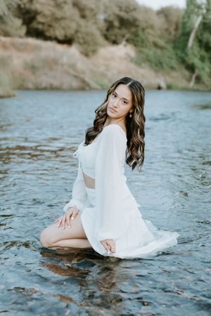 Young woman in white dress sitting gracefully in shallow river water with lush green hillside backdrop.