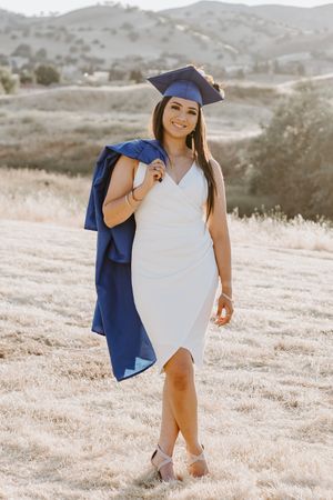 Young woman in white dress and blue graduation cap and gown poses outdoors in a sunny field with rolling hills.