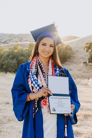 Young woman in blue graduation cap and gown proudly holding diploma outdoors with sunlit hills in background.