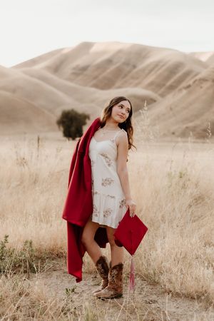 Young woman in floral dress, red blazer, cowboy boots, holding graduation cap in golden field with hills.