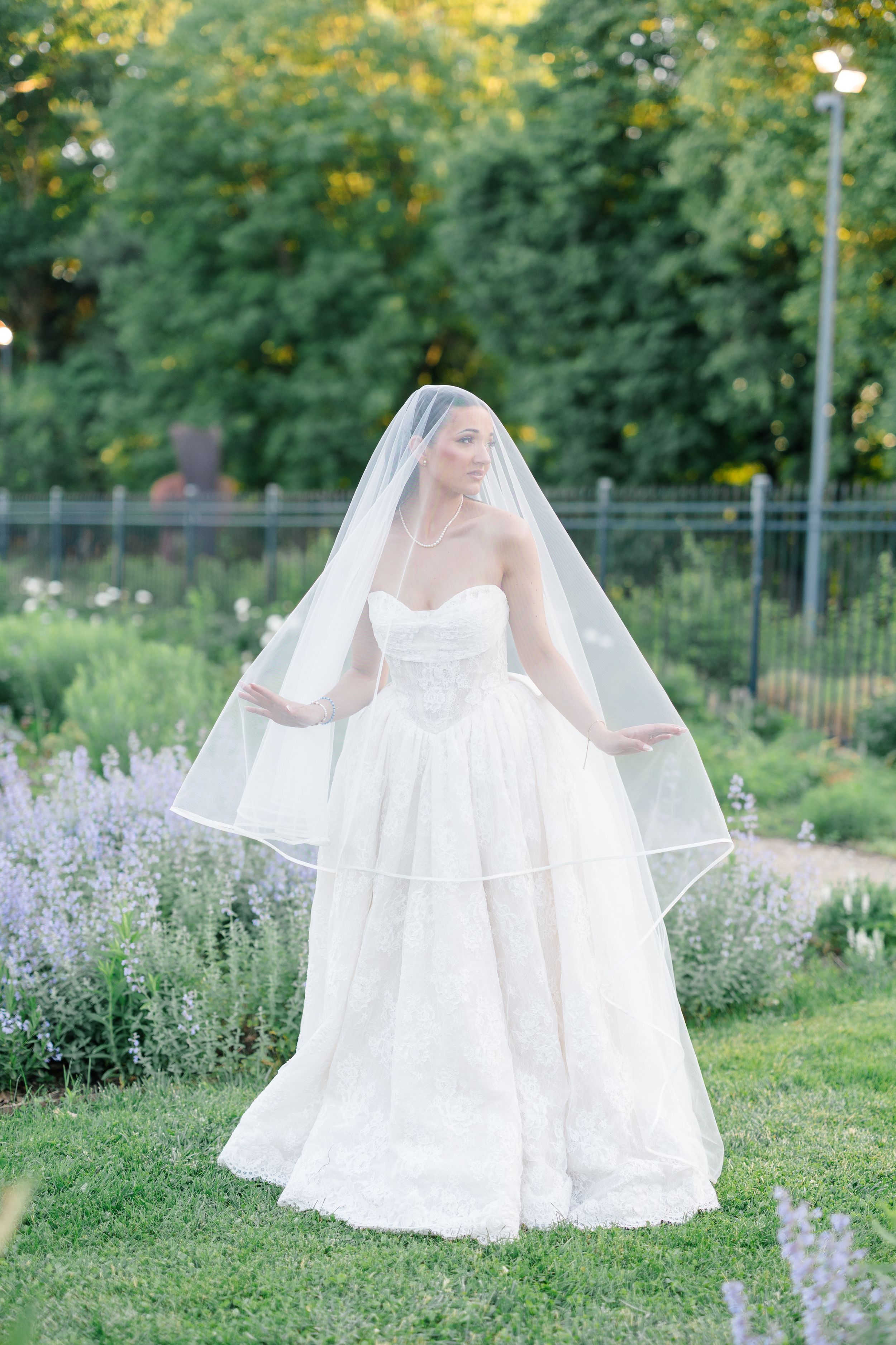 Bride in strapless lace wedding gown and flowing veil poses gracefully in a lush garden with purple lavender flowers.