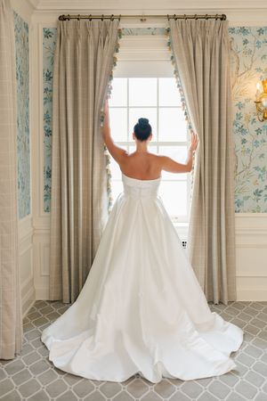 Bride in strapless white ball gown with train stands at window, viewed from behind, in elegant floral wallpapered room.