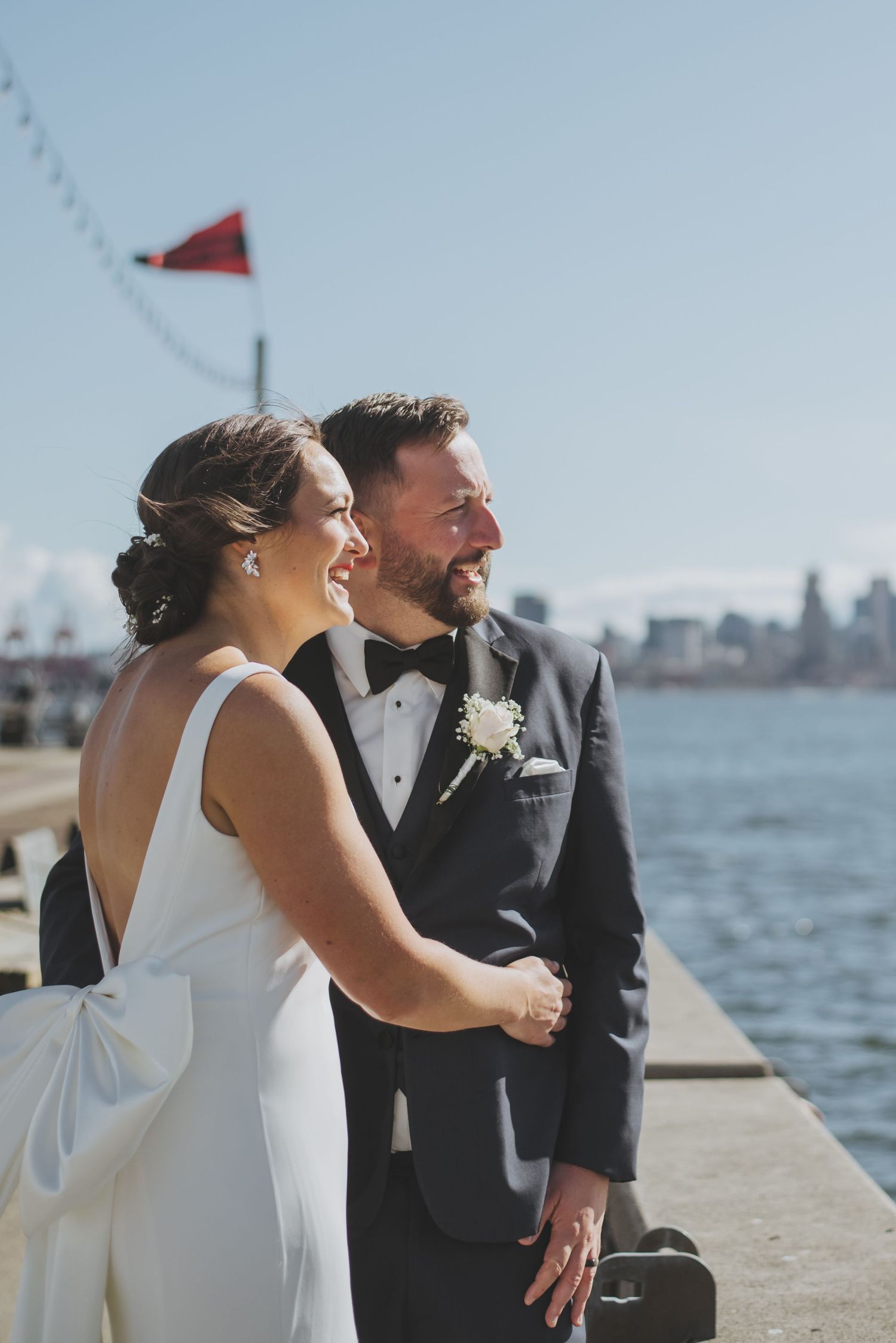 A newlywed couple laughs together on a sunny pier by the water.