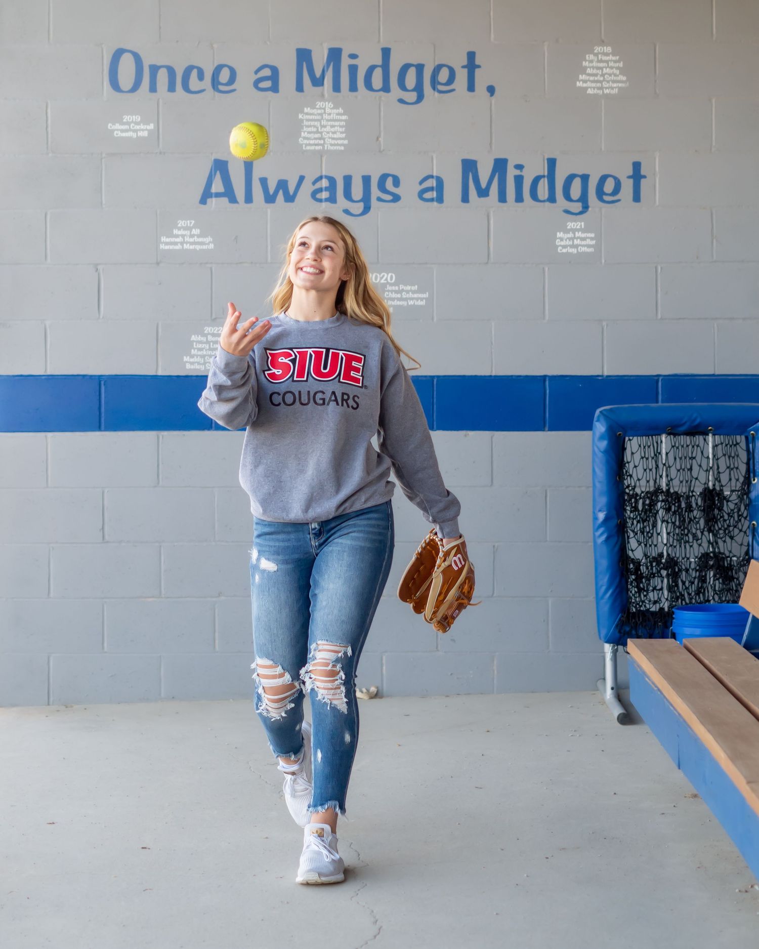 Baseball player in ripped jeans and grey sweater posing in front of blue wall.