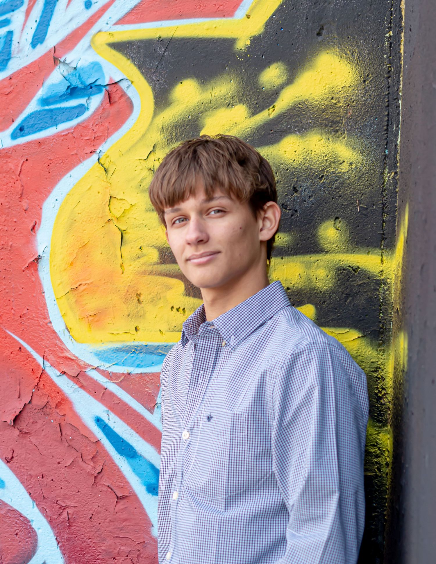 Student poses against colorful graffiti wall wearing blue checkered button-down shirt.