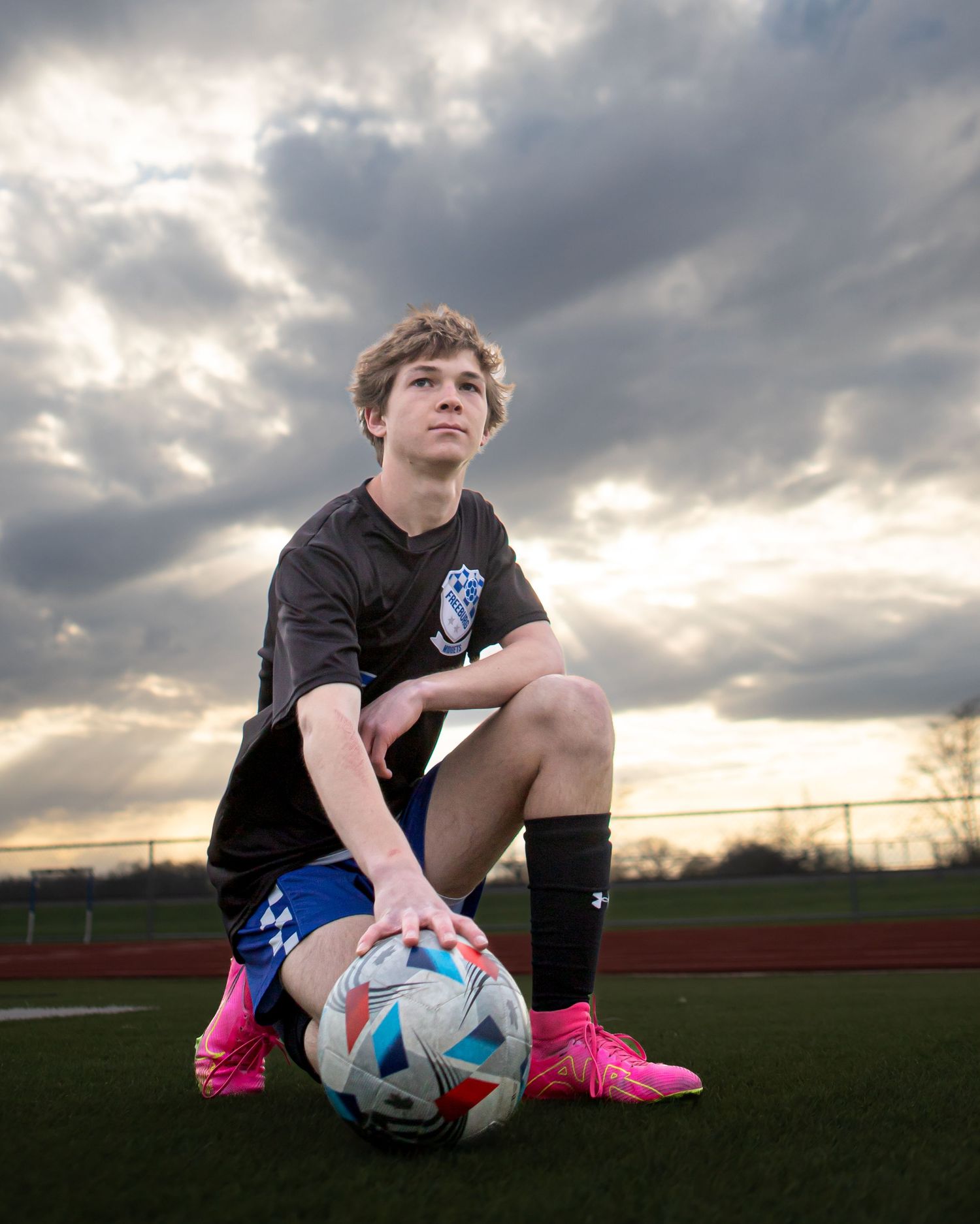 Soccer player sitting with ball on sports field during dramatic sunset.