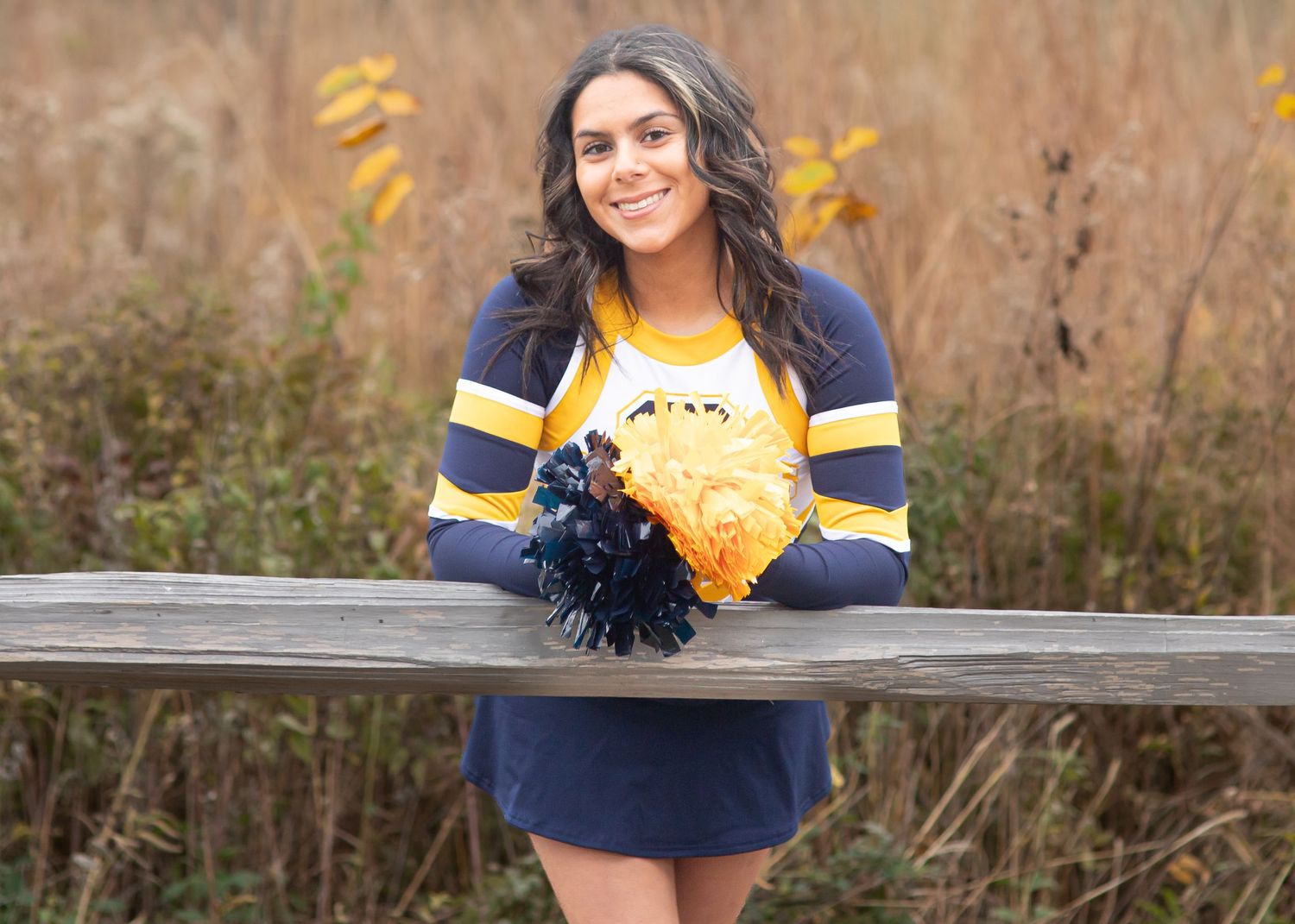 Cheerleader poses in blue and gold uniform against autumn field backdrop.