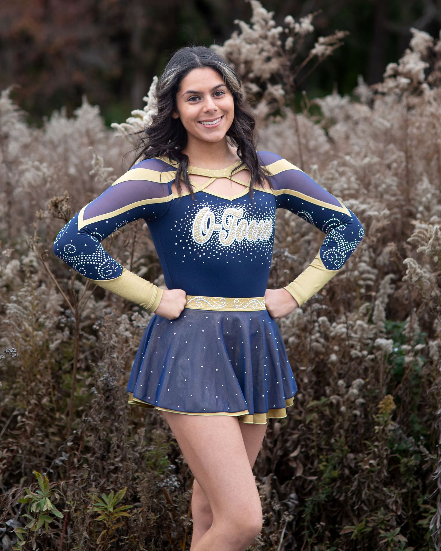 Cheerleader in navy and gold uniform poses with sparkle details against natural backdrop.
