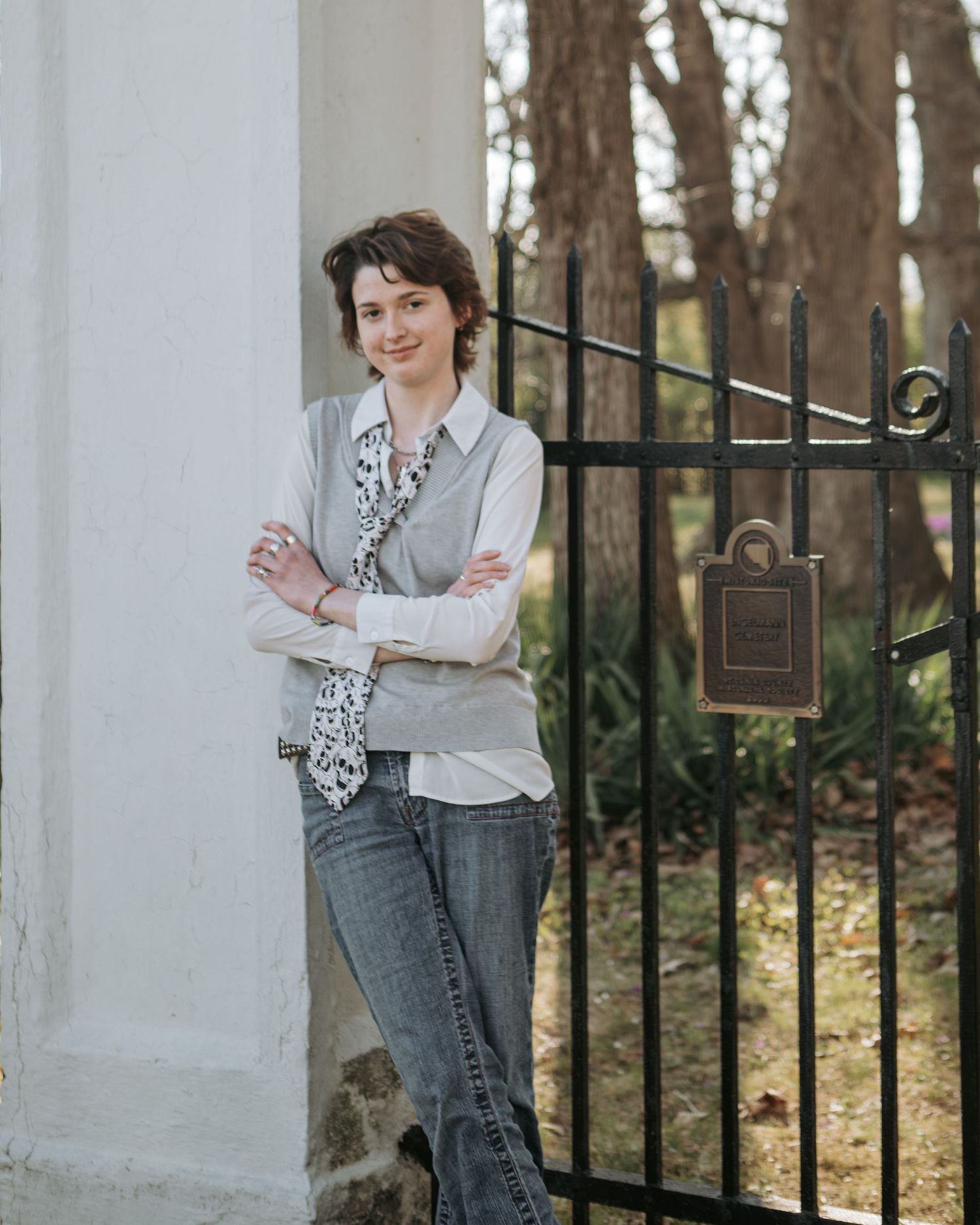 Figure in business casual attire leans against white wall near decorative iron gate.