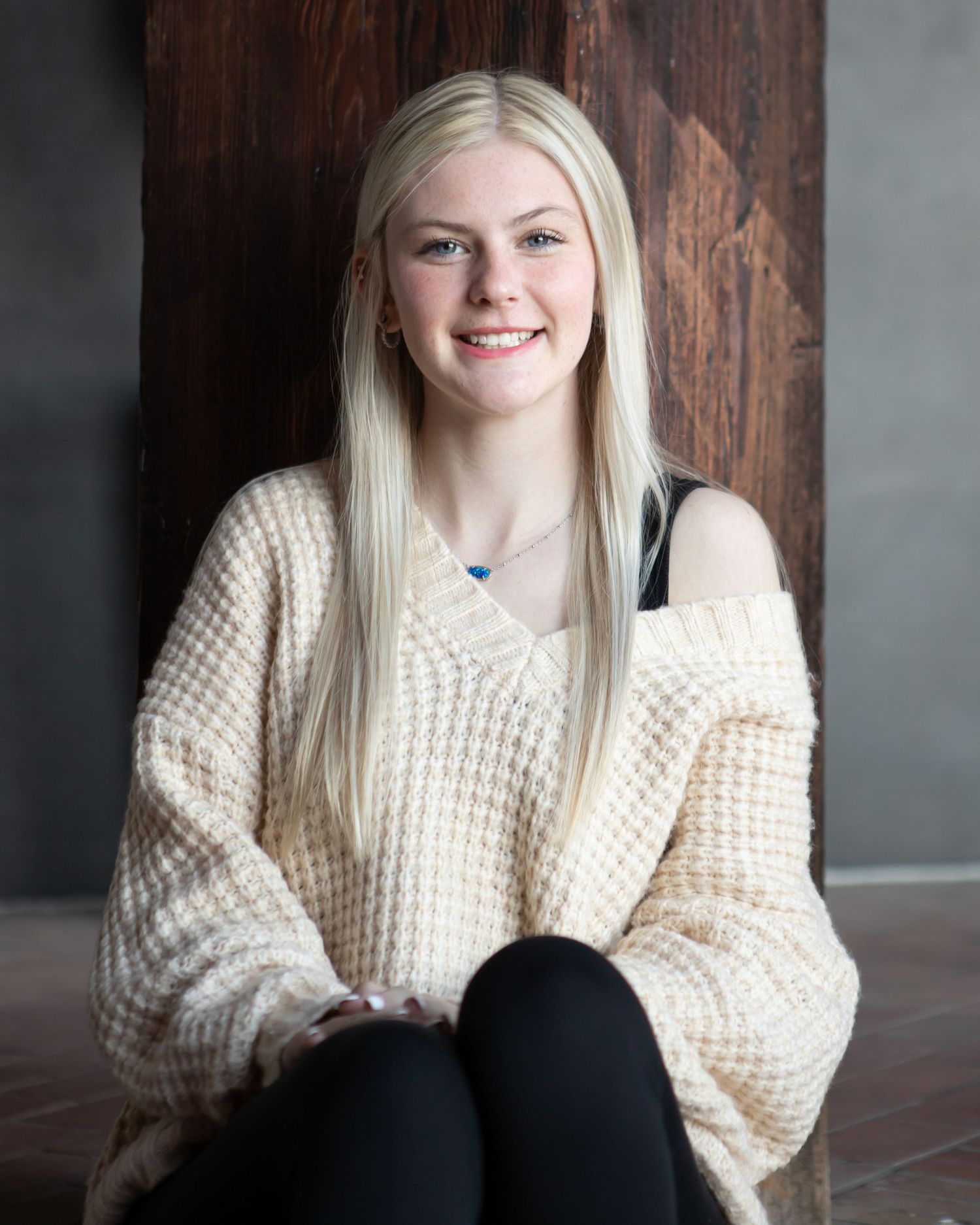 Portrait of someone in cream knit sweater sitting against wooden wall.