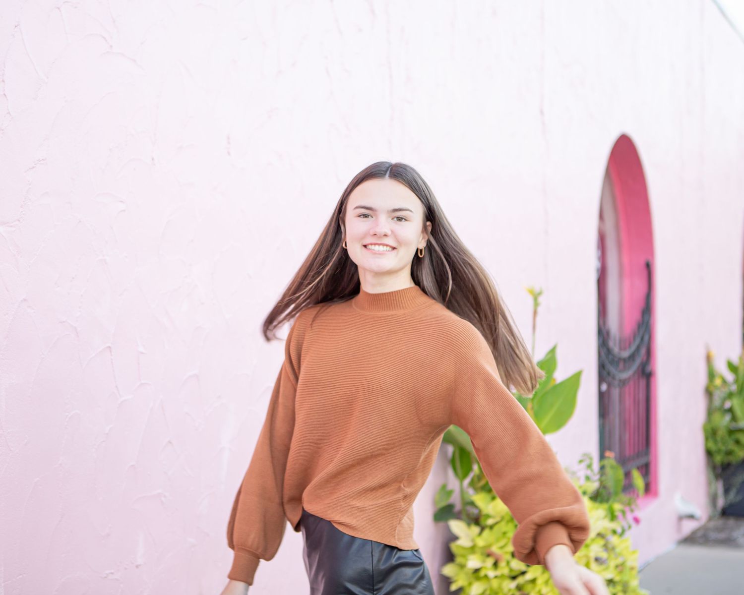 A casual brown sweater outfit is showcased against a pink wall with plants.