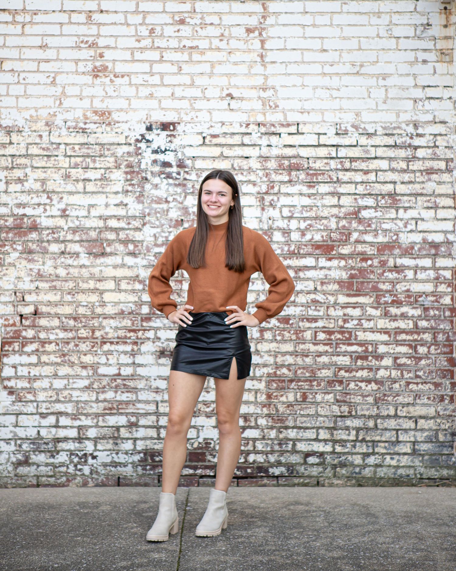 Athletic pose in brown sweater and shorts against a weathered white brick wall.