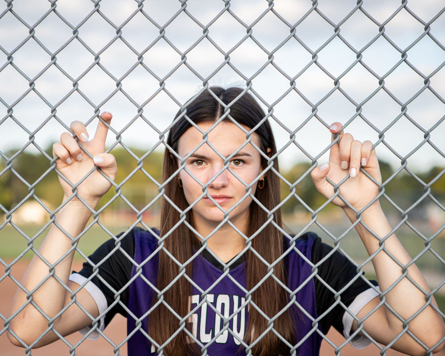 Softball player grips chain link fence while wearing purple team uniform.