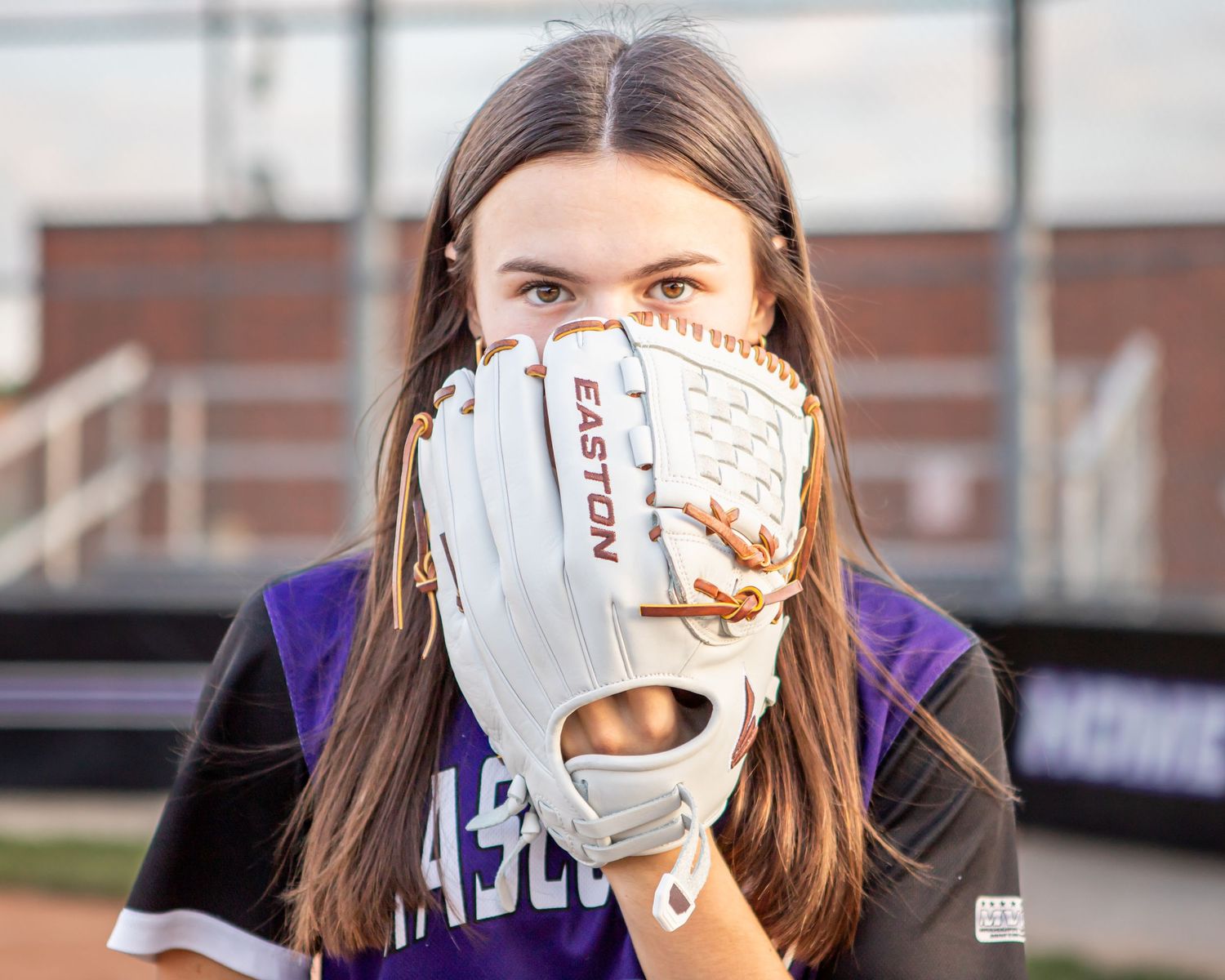 Softball player poses with white glove covering face in purple uniform.