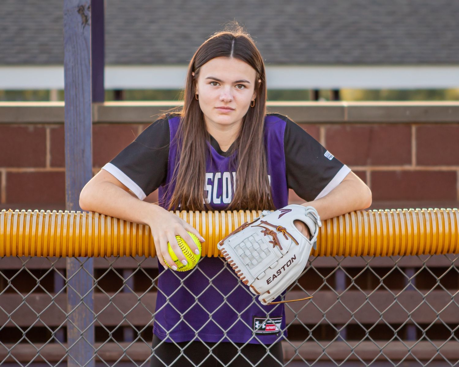 Softball player leans on fence holding yellow ball and glove in purple uniform.