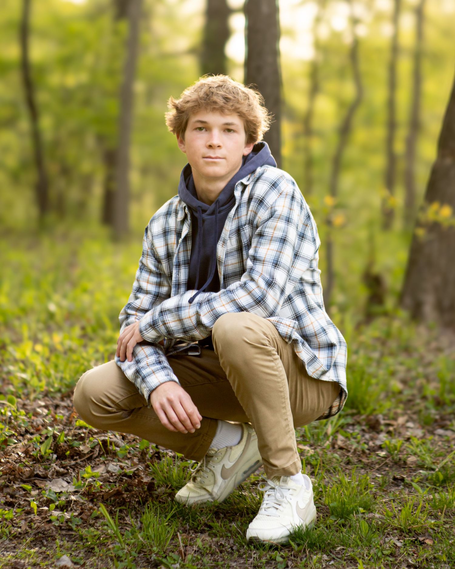 Casual outdoor portrait of person in plaid shirt and khakis crouching in wooded setting.