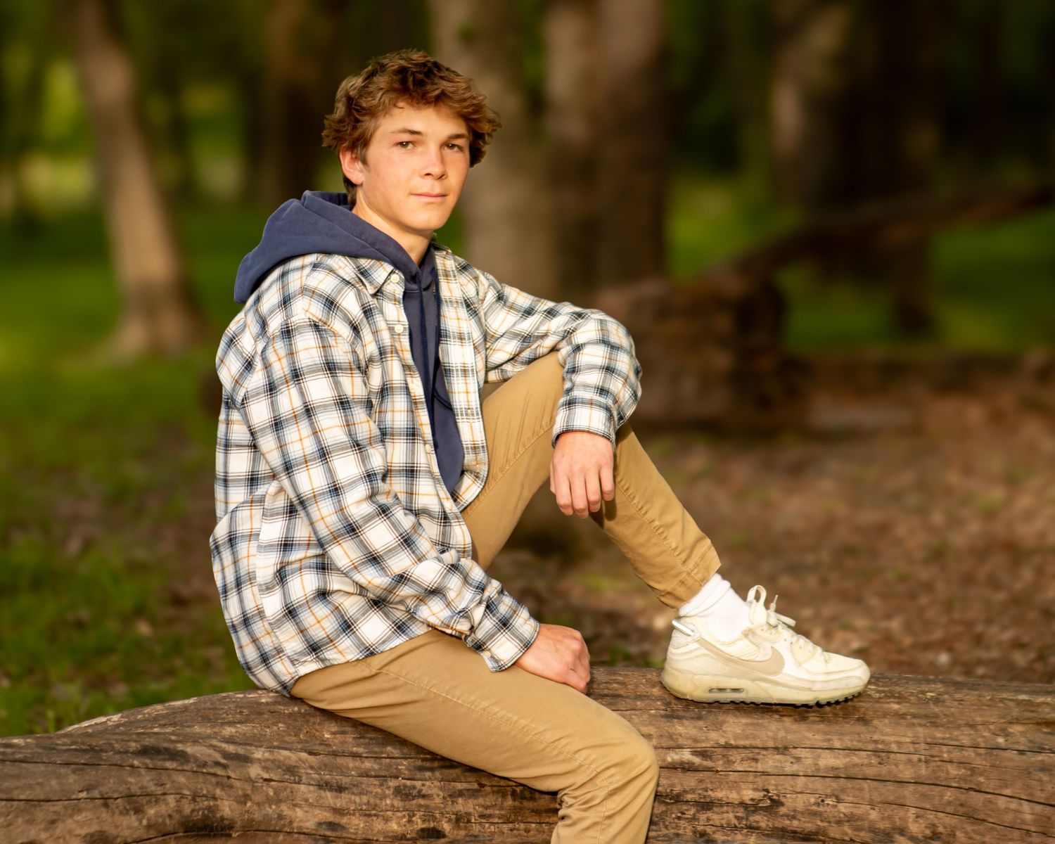 Person in plaid shirt sits casually on log in forest setting.