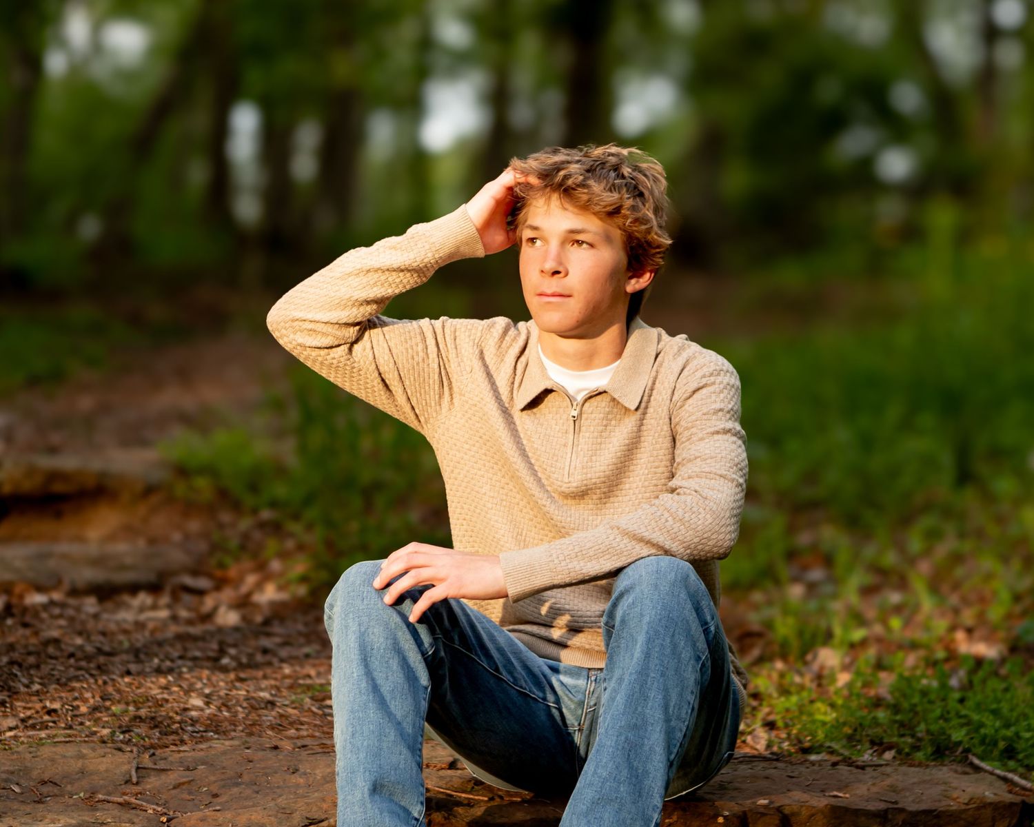 Person in beige sweater sitting on forest path shielding eyes.