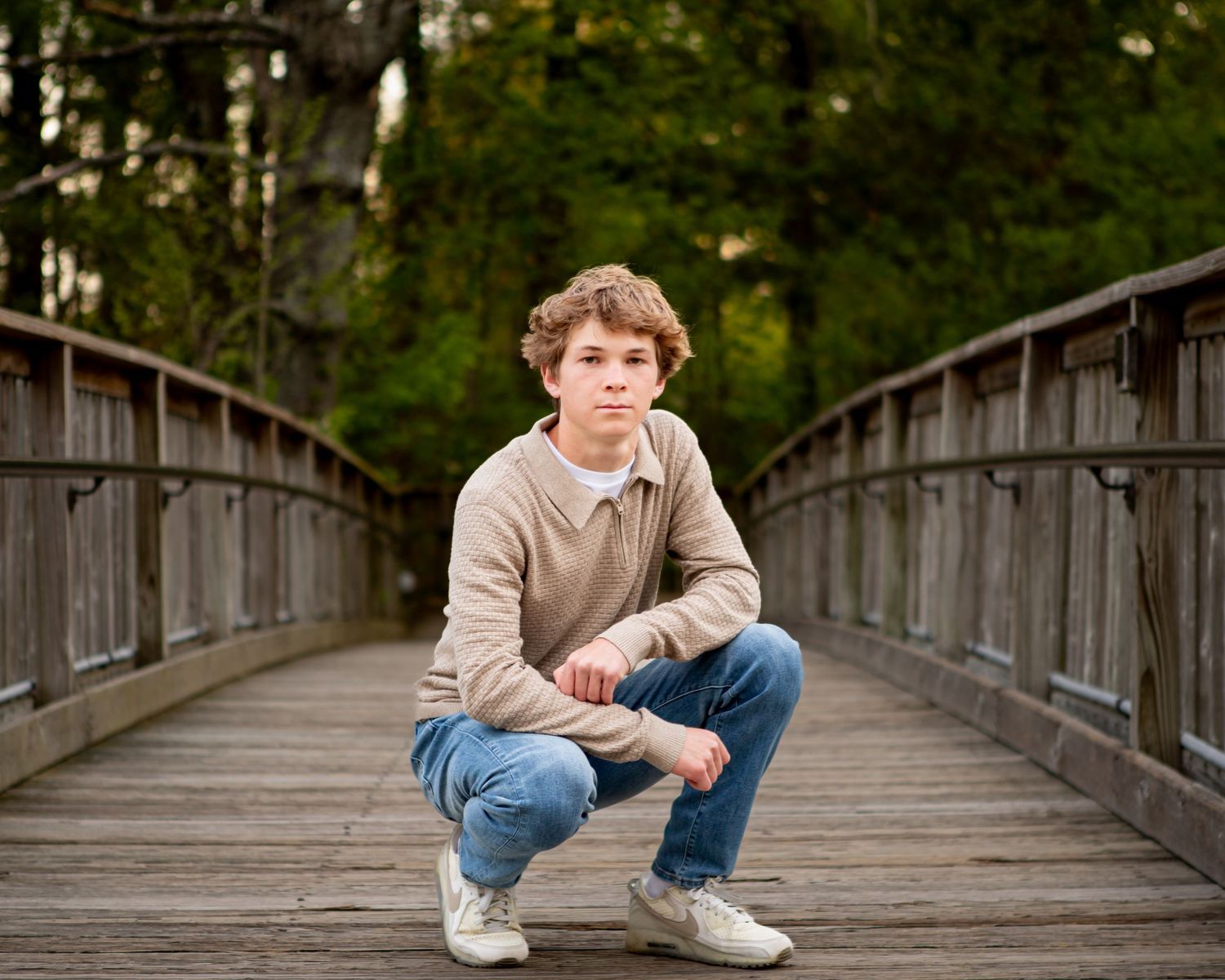 Person crouches on wooden bridge in casual attire surrounded by forest.