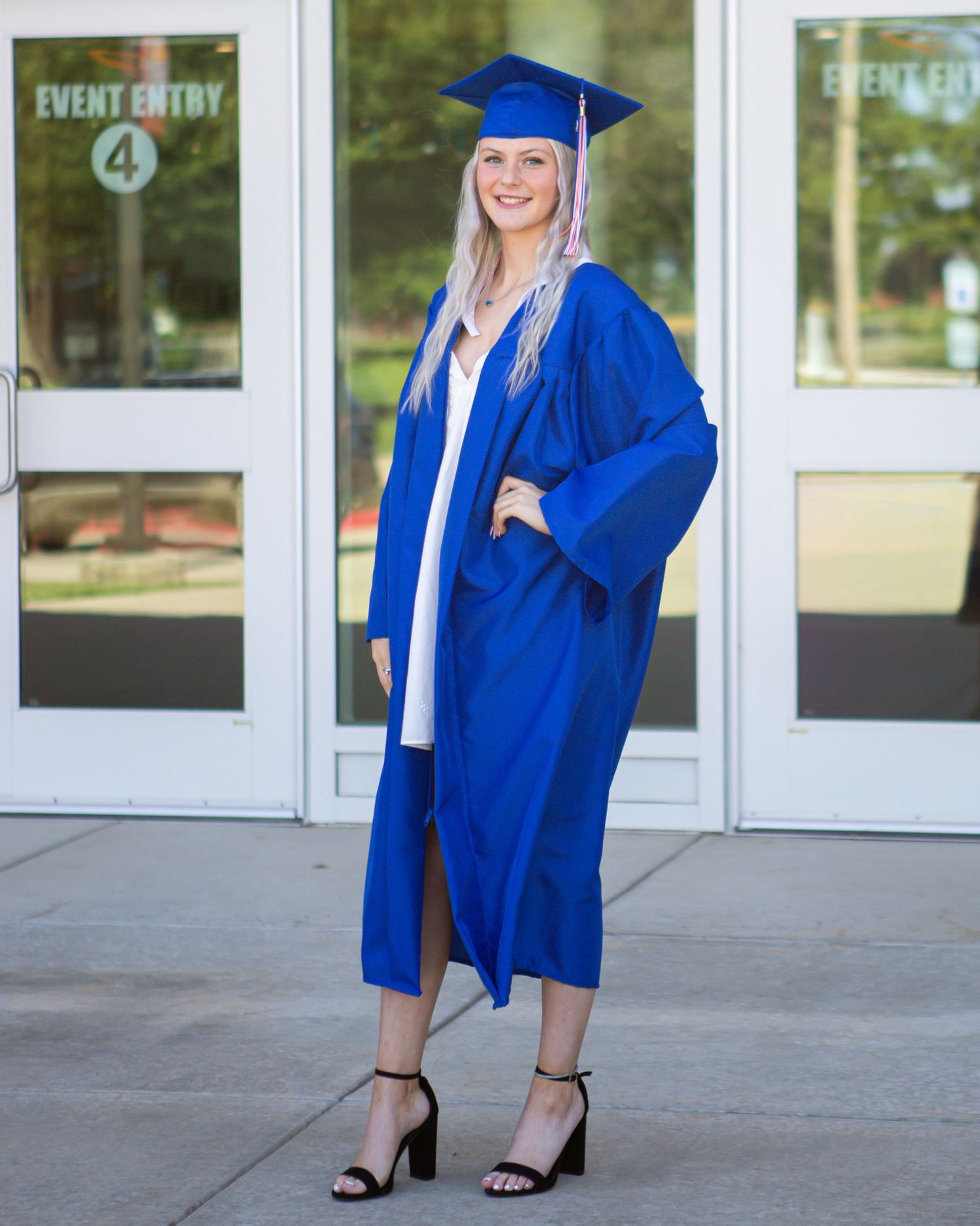 Graduate poses in blue cap and gown outside school entrance doors.