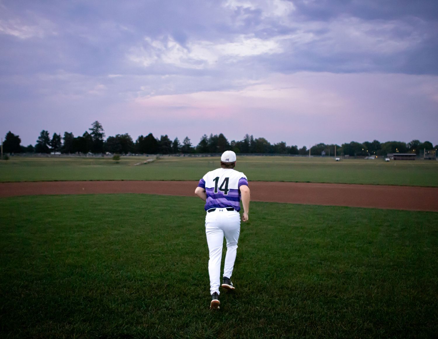 Baseball player walking alone on green field wearing white uniform with number 14.