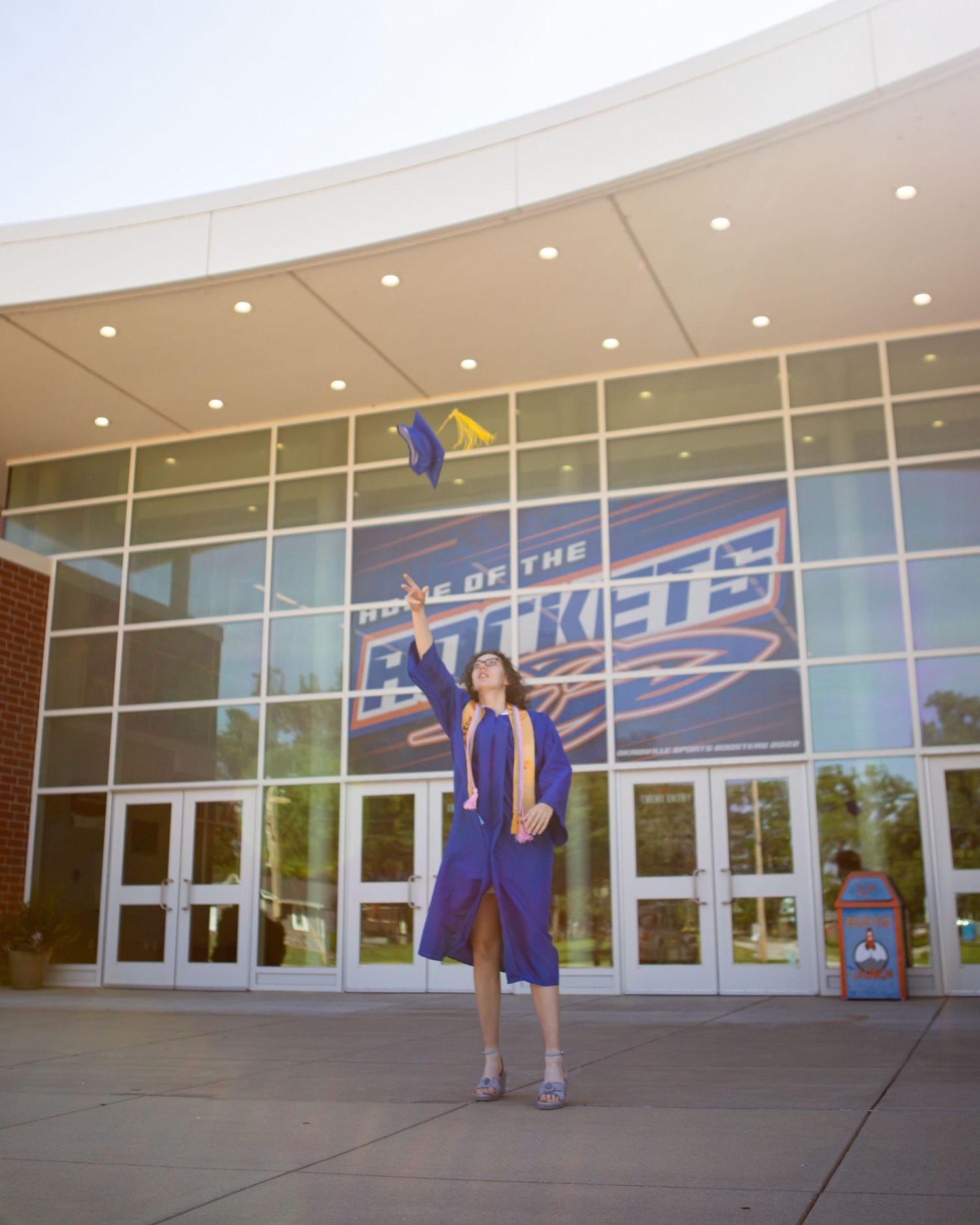 Graduate in blue cap and gown standing in front of Home of the Rockets building entrance.