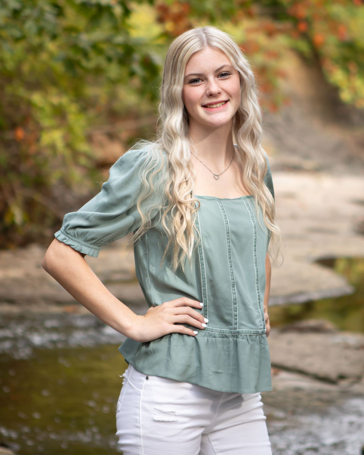 Portrait in sage green top against autumn foliage background near water.