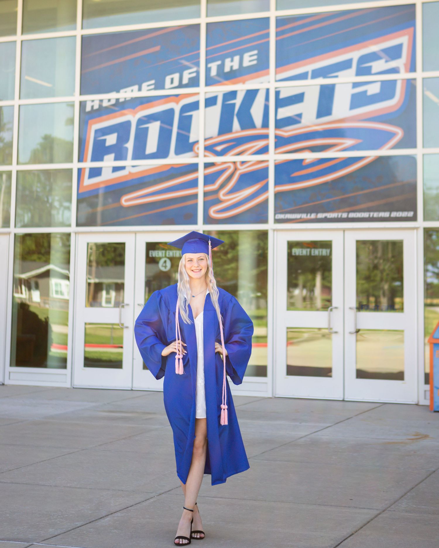 Graduate posing in blue cap and gown in front of Rockets building signage.