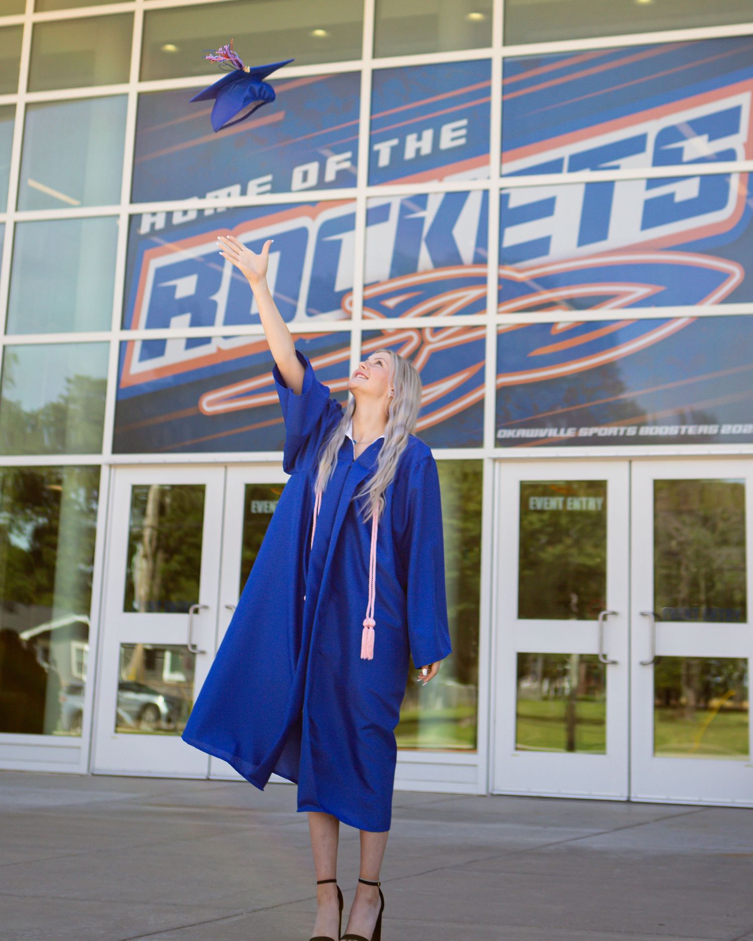 Graduate tossing blue cap in air in front of Rockets building entrance.