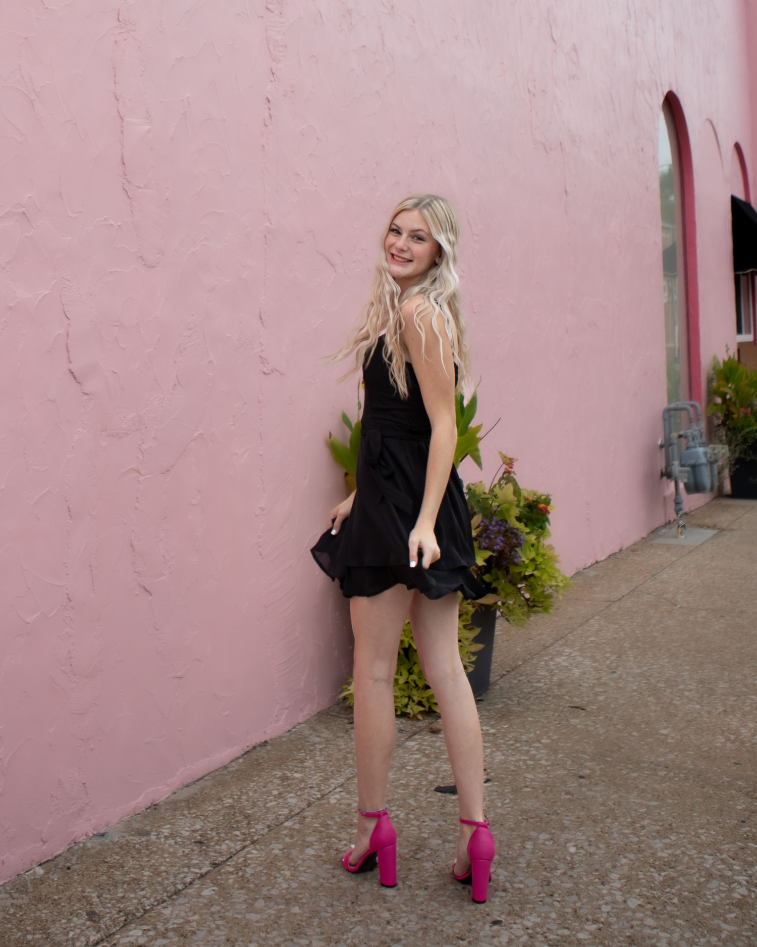 Fashion portrait against pink wall wearing black dress and pink heels.