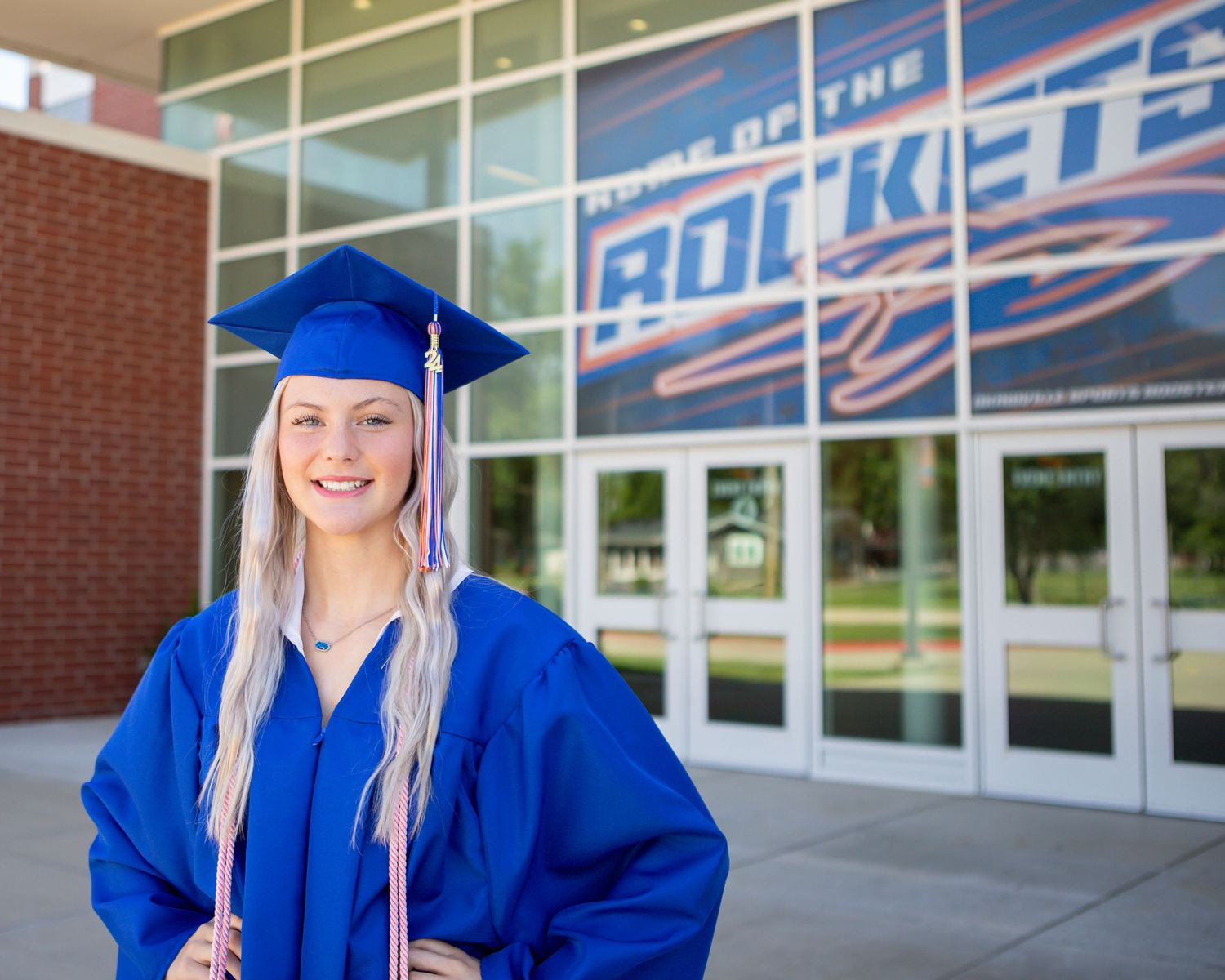 Graduate in blue cap and gown smiling in front of Rockets building entrance.